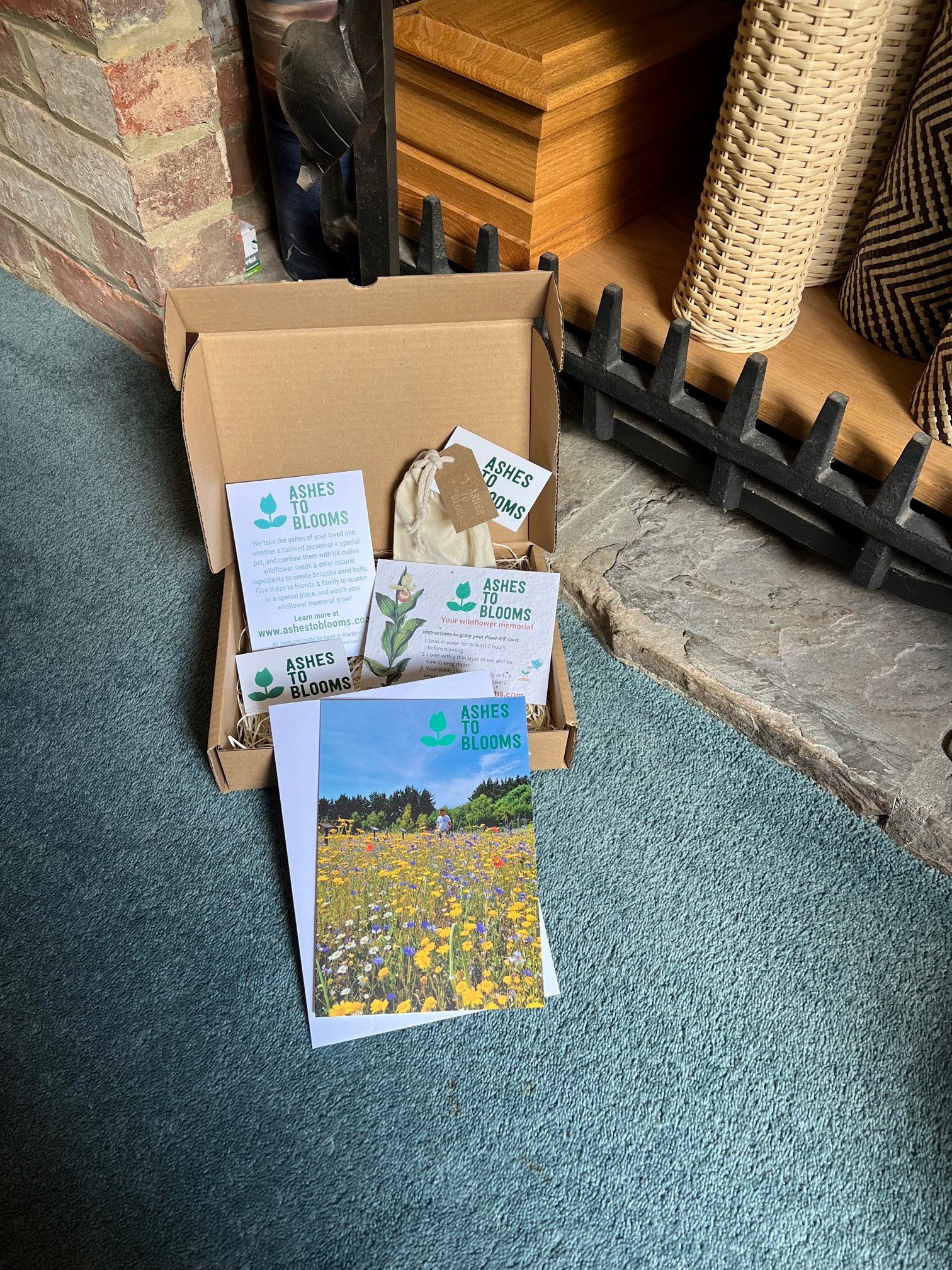 open brown cardboard box containing promotional flyers for Ashes to Blooms sits next to a fireplace. There is also a small cotton bag in the box, and a greetings card depicting a wildflower field in the foreground